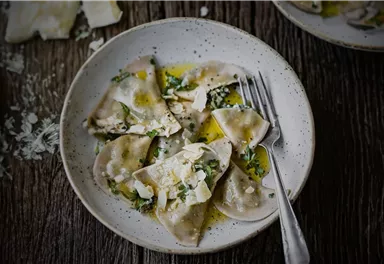 A plate with fresh ravioli, garnished with herbs and a fine oil. Next to it lies a fork on a rustic wooden table.