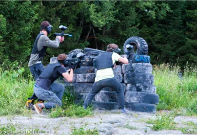 Drei Personen spielen auf einem Outdoor-Gelände mit Paintball-Markierern. Sie nehmen Deckung hinter einer Ansammlung von Reifen und sind von Bäumen umgeben.