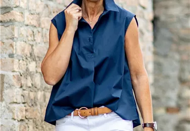 Eine lächelnde Frau trägt ein elegantes, ärmelloses, blaues Hemd und weiße Hose. Sie steht vor einer alten Steinmauer.