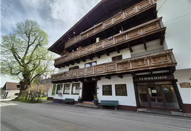 A traditional Alpine building with wooden balustrades and several floors. In front of the house is a quiet street and a large tree.