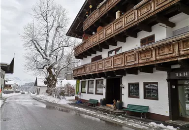 A snowy street with a traditional alpine building. In the background, there is a large tree without leaves.