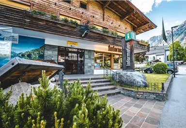 Ein informatives Gebäude aus Holz mit der Aufschrift „Information“ an einer malerischen Straße. Im Vordergrund sind grüne Pflanzen und ein gemütliches Ambiente sichtbar.