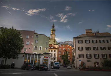 A historical cityscape with colorful buildings and a distinct church tower. Bright clouds drift across the sky while trees stand in the foreground.