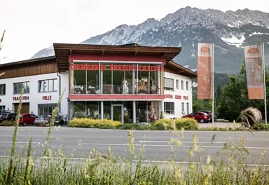 Ein modernes Gebäude mit großen Fenstern und einem klaren Blick auf die umliegenden Berge. Im Vordergrund sind Fahrzeuge und Grünflächen zu sehen.
