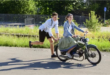 Ein Junge schiebt ein Mädchen, das auf einem Motorrad sitzt, während sie auf einer Straße fahren. Es ist ein sonniger Tag mit viel Grün im Hintergrund.