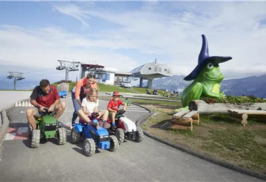 A family is riding on small tractors along a track. In the background, there is a large green figure of a frog wearing a hat.