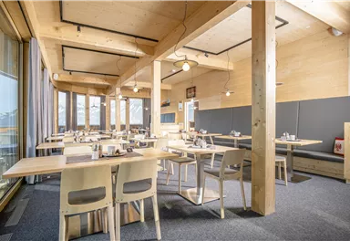 A modern restaurant with wooden elements and large windows. The tables are neatly set and offer space for guests.