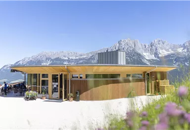 A modern building in the mountains with wooden cladding. In the background, impressive mountain peaks and a clear blue sky can be seen.