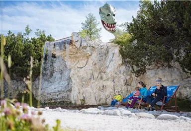A family sits relaxed on deck chairs in nature. In the background, a huge, fantastical dragon can be seen.