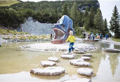 A child jumps on stones in a water basin with a large fish sculpture in the background. In the background, more visitors and trees can be seen.