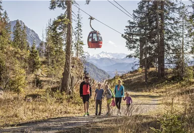 A family is hiking on a path through the mountains. A cable car is gliding above them in the clear sky.