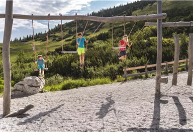 A playground scene with three children playing on swings. In the background, green meadows and hills can be seen.