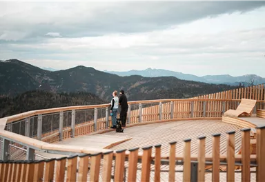 A viewpoint in the mountains with a wooden railing. Two people are enjoying the view of the hilly landscape under a cloudy sky.