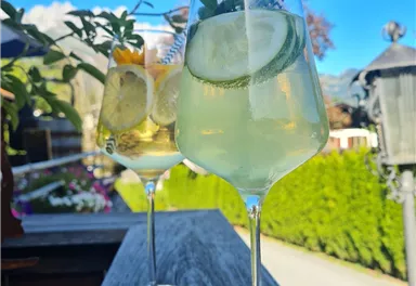 Two large glasses with refreshing cocktails are placed on a wooden railing. In the background, the blue sky and green landscape shine.