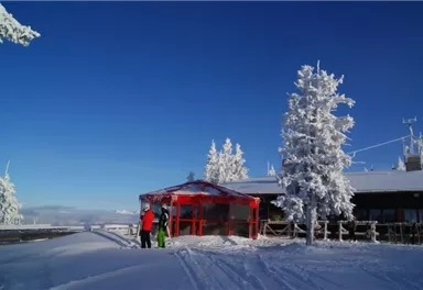 A snow-covered landscape with a red building and tall, snow-covered trees. In the foreground, two people are standing in ski clothing.