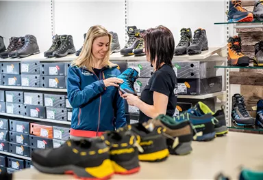 A customer is trying on a blue shoe while a saleswoman helps her. In the background, there are many shoes in different colors and styles visible.