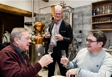 Three men toast with glasses while standing in a distillery. In the background, equipment for producing spirits is visible.