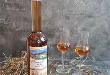 Eine Flasche Zwetschgenwasser steht auf einem grauen Hintergrund, umgeben von Heu. Neben der Flasche stehen zwei kleine Gläser mit dem klaren, goldenen Getränk.