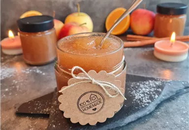 Ein Glas Apfelmus mit Löffel, dekoriert mit einem Etikett, steht auf einem schwarzen Teller. Im Hintergrund sind Äpfel und Kerzen zu sehen, die eine gemütliche Atmosphäre schaffen.