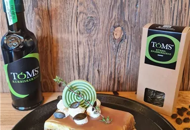 Ein Stück leckere Torte auf einem schwarzen Teller, dekoriert mit Sahne und einer grünen Ringdekoration. Daneben stehen eine Flasche und eine Verpackung von TÖM'S Kräuterlikör.