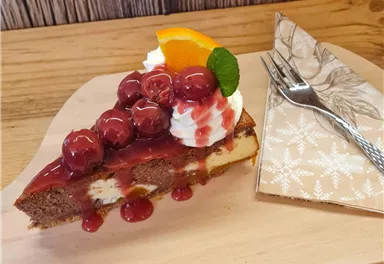 Ein Stück Käsekuchen mit Kirschen, Sahne und einer Orangenscheibe. Das Dessert liegt auf einem Holzbrett neben einer Gabel.