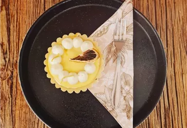 Ein köstlicher Zitronenkuchen mit einer dekorativen Sahnehaube auf einem schwarzen Teller. Daneben liegt eine Serviette und eine Gabel.