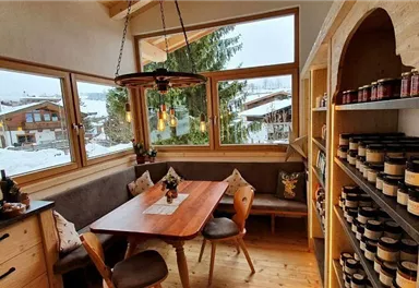 Ein gemütlicher Raum mit großen Fenstern, der einen Blick auf verschneite Häuser und Bäume bietet. Am Tisch stehen Holzstühle und eine dekorierte Tischdecke, während Regale mit verschiedenen Produkten gefüllt sind.