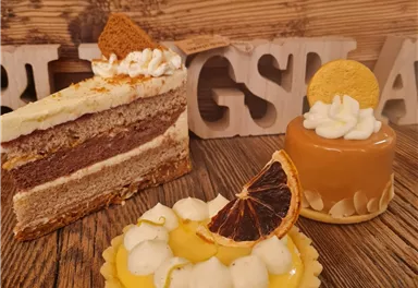 Three delicious desserts are on a wooden table. A layer cake, a small caramel pastry, and a lemon tart with cream and decoration.