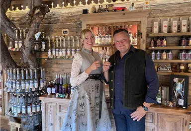 A couple stands at a wooden counter in a traditional shop. They are holding glasses in their hands and smiling kindly.