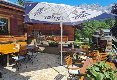 An inviting terrace with wooden chairs and tables, shaded by a large umbrella. In the background, green trees and mountains can be seen.