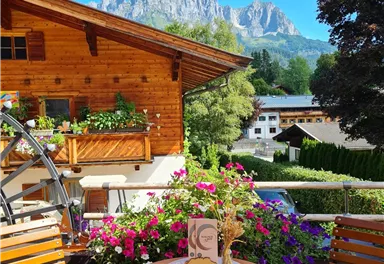 A cozy balcony with a round table and chairs. In the background, you can see charming mountains and blooming flowers.