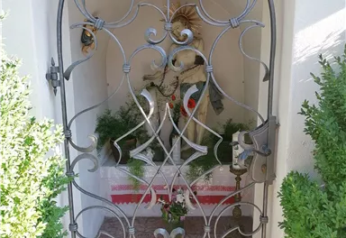 A wrought-iron gate leads to a niche with religious ornaments. Plants frame the area, giving it a serene atmosphere.