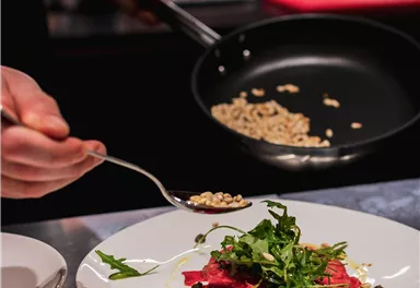 Ein Koch präsentiert einen Teller mit dünn geschnittenem Fleisch, garniert mit frischen Kräutern. Im Hintergrund wird eine Zutat aus einer Pfanne auf den Teller gestreut.