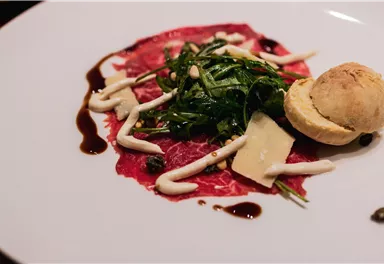 Ein hübsches Gericht mit dünn geschnittenem Fleisch, frischem Blattspinat und einem kleinen Brötchen. Die Anrichtung ist geschmackvoll mit einer cremigen Sauce und Balsamico-Reduktion.