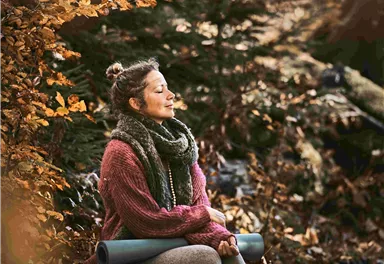 A woman is sitting amidst autumn leaves and enjoying nature. She is wearing a warm sweater and has a yoga mat with her.