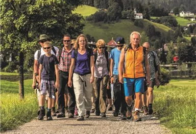 Eine Gruppe von Wanderern spaziert einen schmalen Weg entlang, umgeben von grünen Hügeln. Die Teilnehmer tragen Wanderbekleidung und scheinen die Natur zu genießen.