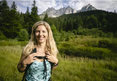 A cheerful woman stands in a green landscape with mountains in the background. She is wearing a backpack and smiling.