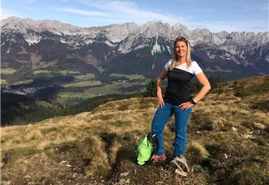 A woman stands on a mountain with a breathtaking view of the Alps. In the background, snow-covered peaks and green valleys can be seen.