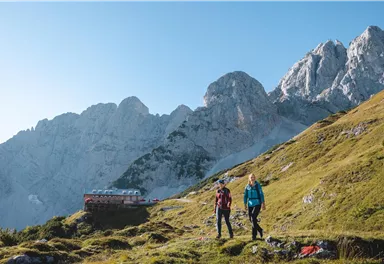 Zwei Wanderer stehen auf einer grünen Wiese in den Bergen. Im Hintergrund sind majestätische Felsen und eine Berghütte zu sehen.
