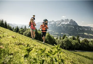 Eine Familie wandert mit Rucksäcken auf einem grünen Hügel. Im Hintergrund sind majestätische Berge und ein klarer Himmel zu sehen.