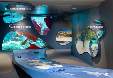 A modern exhibition space with interactive displays and blue accents. The walls feature underwater-like scenes and information about water ecosystems.
