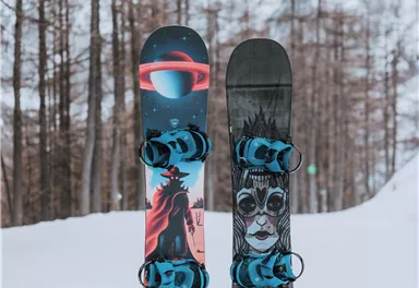 Two snowboards stand in the snow, surrounded by snow-covered trees. The left board features a colorful design, while the right board displays a graphic face.