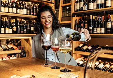 A smiling woman pours wine into three glasses. In the background, numerous wine bottles are visible.
