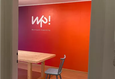 Ein freundlicher Coworking-Raum mit einem großen Tisch und einem Stuhl. Die Wand ist in warmen Farben gestaltet und trägt das Logo „wp!“.