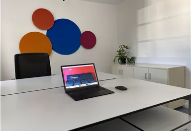 Ein modernes Büro mit einem großen weißen Tisch und einem Laptop. An der Wand sind bunte Kreise und eine Pflanze ist sichtbar.