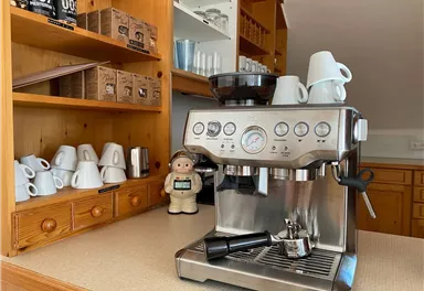 Eine moderne Kaffeemaschine steht auf einer Arbeitsfläche. Im Hintergrund sind verschiedene Kaffeetassen und eine Dekorationsfigur sichtbar.