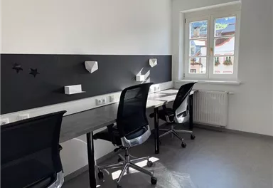 Ein modernes Büro mit hellen Wänden und großen Fenstern. Es gibt mehrere Schreibtische mit Stühlen und eine minimalistische Gestaltung.