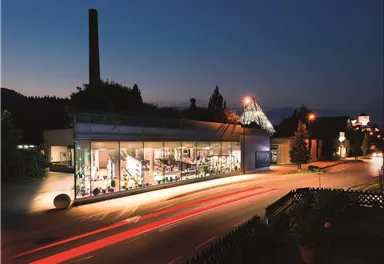 A modern building with large windows that is illuminated at night. In the foreground, there are streetlights and plants visible.
