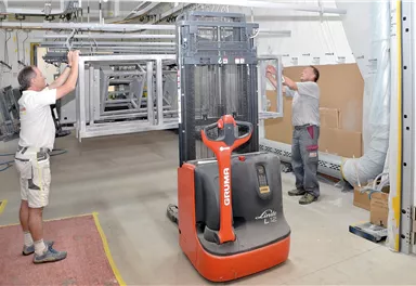 Two workers in a warehouse are assembling frame elements. In the foreground, a forklift is standing.