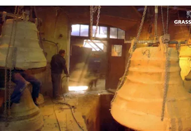 In a workshop, large bells hang from the ceiling. A worker is checking the surroundings.